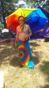 Rainbow Bloc Kit is dressed in a rainbow sarong, rainbow face bandanna, and holding a rainbow umbrella while standing outside the Purple Taco.
