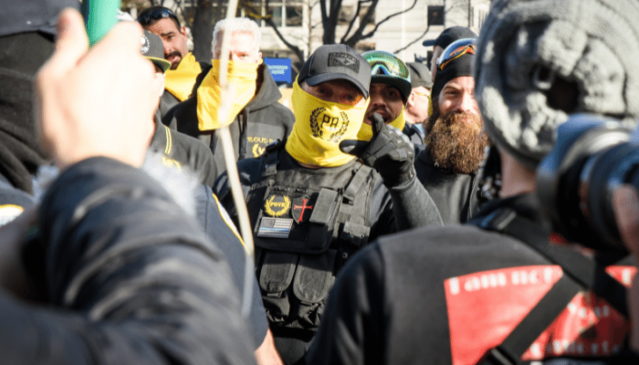 In a heated confrontation, Proud Boys in their yellow uniforms confront a group facing away from the camera. One in a yellow Proud Boys mask points angrily at us.