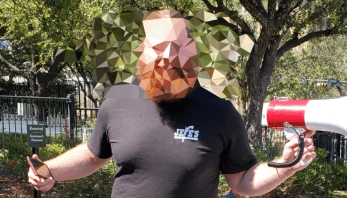 A bald man with a red beard in a black tee gestures, phone in one hand, megaphone in the other, outside the Texas Governor's Mansion. His face has been mosaic blurred.