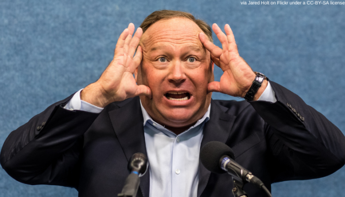 Speaking at some microphones on a podium, Alex jones holds his hands to either side of his head as he makes an agitated, yelling expression. Added text: via Jared Holt on Flickr under a CC-BY-SA license