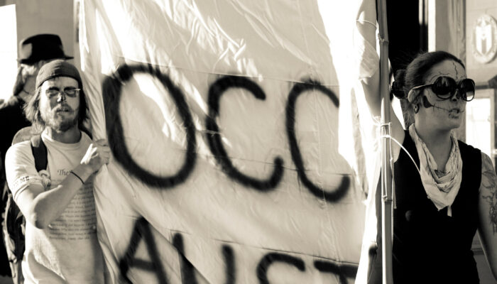 Two activists in zombie facepaint march while carrying a simple Occupy Austin banner made with black spraypaint on a white sheet.