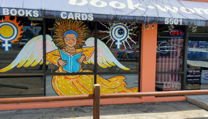 The storefront for Bookwoman in Austin, Texas shows an angel with a halo of curly brown hair, reading a book, her wings outstretched and wearing a golden dress with white stars. She's flanked on either side by female symbols which are in flames or silver electricity.