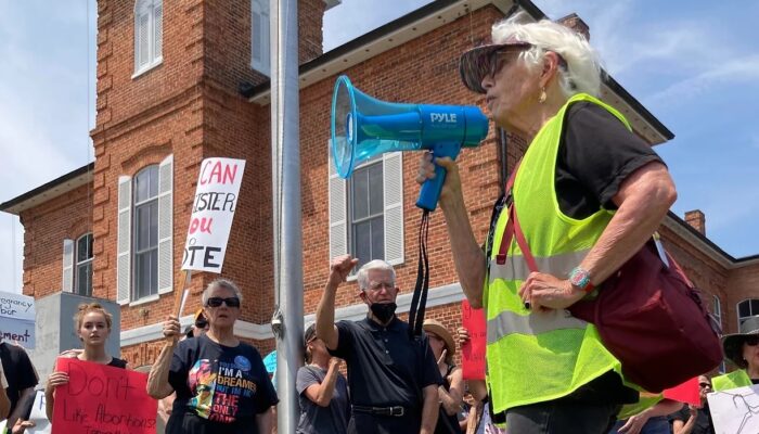 Rev. Carter Heyward, an older white woman with white hair, speaks into a bullhorn, wearing a safety vest, with a rally for abortion access behind her.