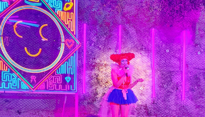 Photo of on stage at Cheer Up Charlie's during Democrasexy's "Carnival of Care", with a huge multi-colored neon smiley face sign behind her. Becky is wearing an outrageous clown outfit, including white boots, a frilly blue miniskirt, orange top and a bright red clown wig shaped into two large buns. The stage is bathed in purple light.