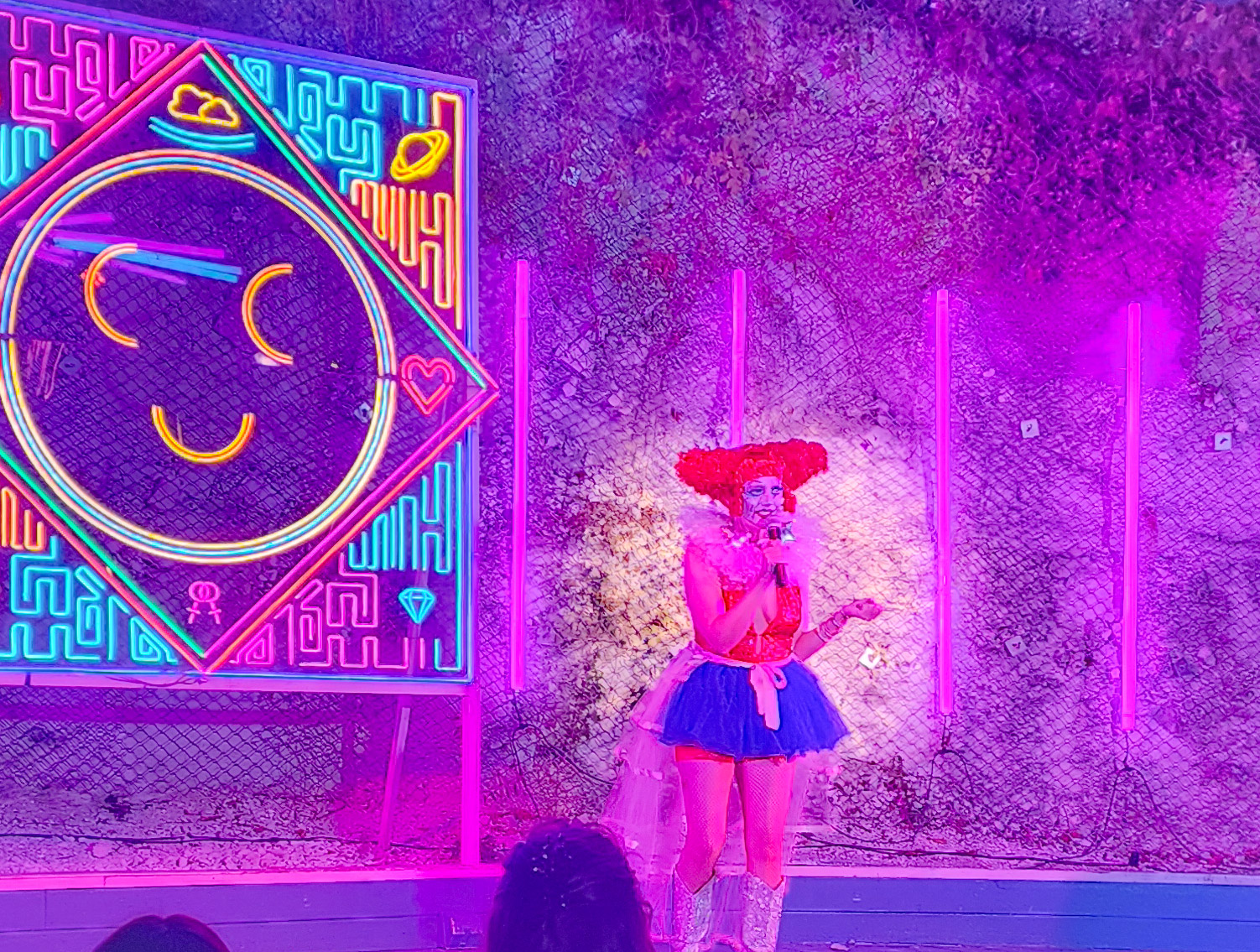 Photo of on stage at Cheer Up Charlie's during Democrasexy's "Carnival of Care", with a huge multi-colored neon smiley face sign behind her. Becky is wearing an outrageous clown outfit, including white boots, a frilly blue miniskirt, orange top and a bright red clown wig shaped into two large buns. The stage is bathed in purple light.