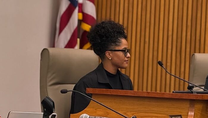 Amanda Rodriguez wearing a dark suit jacket over a dark top, is sitting behind the dais of the San Marcos City council, looking to the right of the camera.