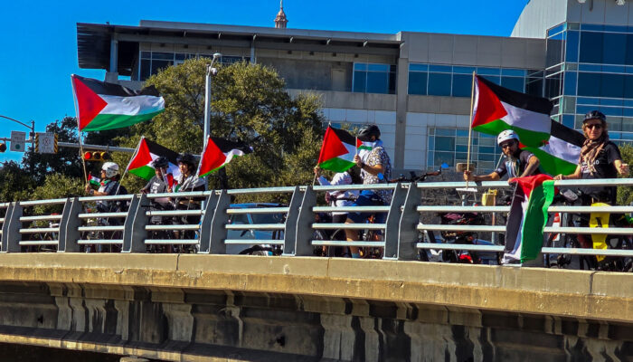 Cyclists on a highway overpass in Austin, Texas hold Palestinian flags which blow in the breeze under a blue sky.
