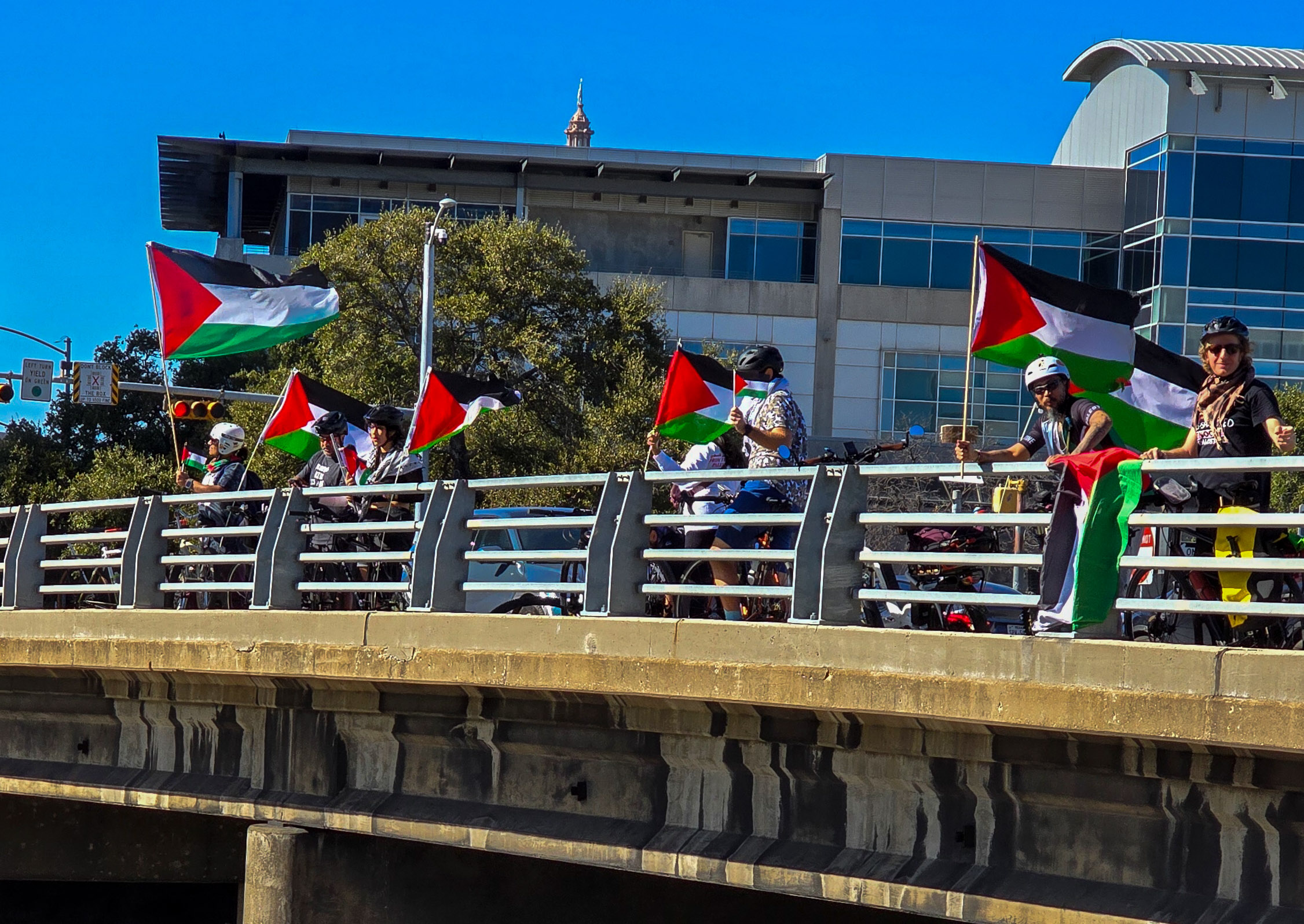 Cyclists on a highway overpass in Austin, Texas hold Palestinian flags which blow in the breeze under a blue sky.