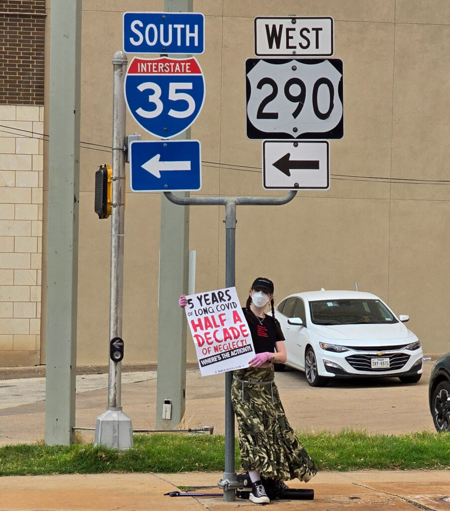 KD Drackert stands under a pair of interstate signs near an Austin highway frontage road, holding a sign which reads: "5 Years of Long Covid, Half a Decade of Neglect! Where's the Action?!" They are wearing a black baseball cap, white face mask, black tee, an ankle-length patterned green skirt blowing in the wind, over low black Converse style sneaker-boots. Their hair falls in two long braids.