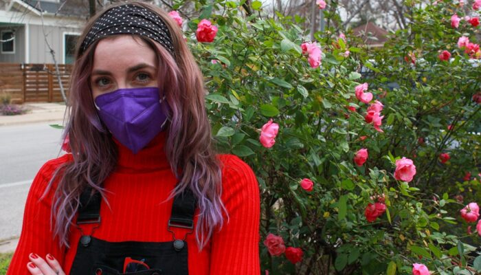 KD Drackert poses with their arms crossed in front of a rose bush in bloom on a suburban street. They are looking directly at the camera over their purple facemask. Their wavy hair has colorful purple highlights. KD is wearing black overalls over a red sweater which matches her manicure.