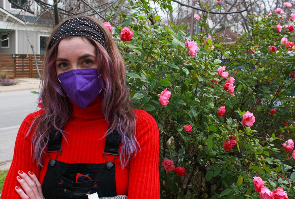 KD Drackert poses with their arms crossed in front of a rose bush in bloom on a suburban street. They are looking directly at the camera over their purple facemask. Their wavy hair has colorful purple highlights. KD is wearing black overalls over a red sweater which matches her manicure.