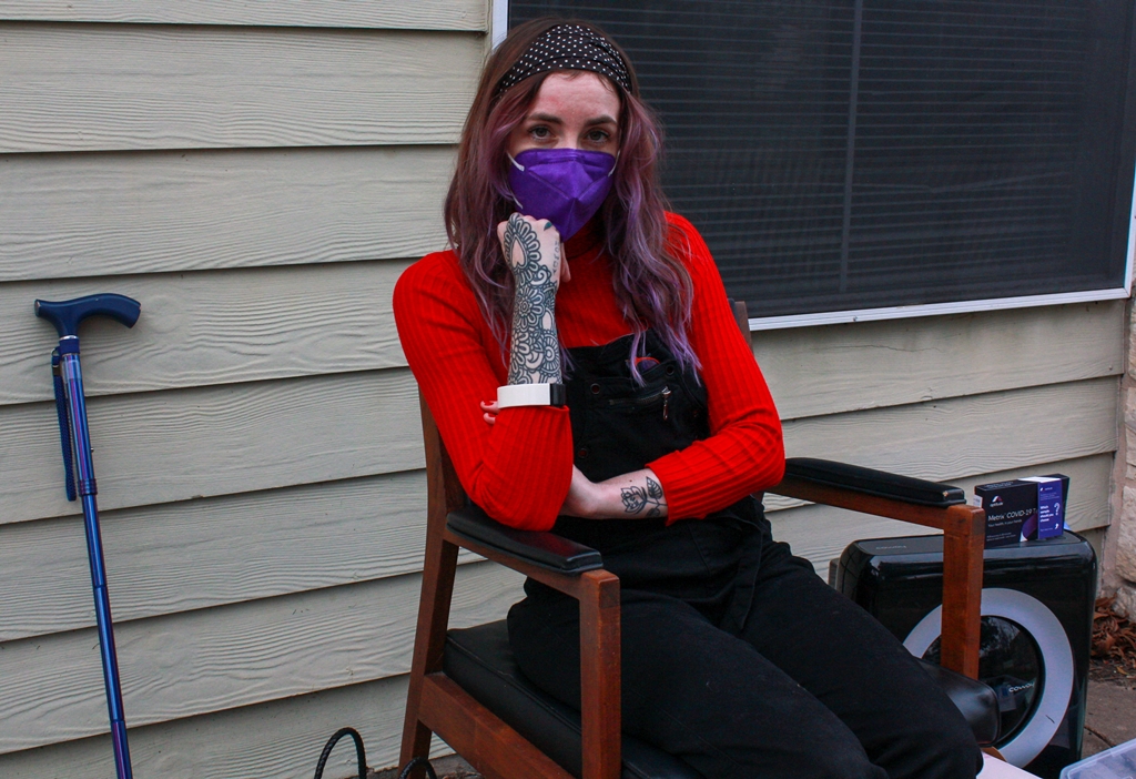 KD is wearing the same outfit as the previous photo, but is now sitting on a chair in a backyard of a home, surrounded by the devices she uses to support their health as a long hauler, including a purple expanding cane and an air purifier. They are resting their chin on their fist, and their sleeves have pulled back partly to reveal dense tattoos on their arms. 