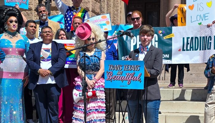 Ash Hall is a nonbinary white person with short brown hair in a pageboy cut, wearing a suit with a trans pride bowtie, speaks at a podium labeled Trans Texans are Beautiful. Other allies are arrayed behind them including colorful drag queens like Brigitte Bandit and Maxine LaQueen.