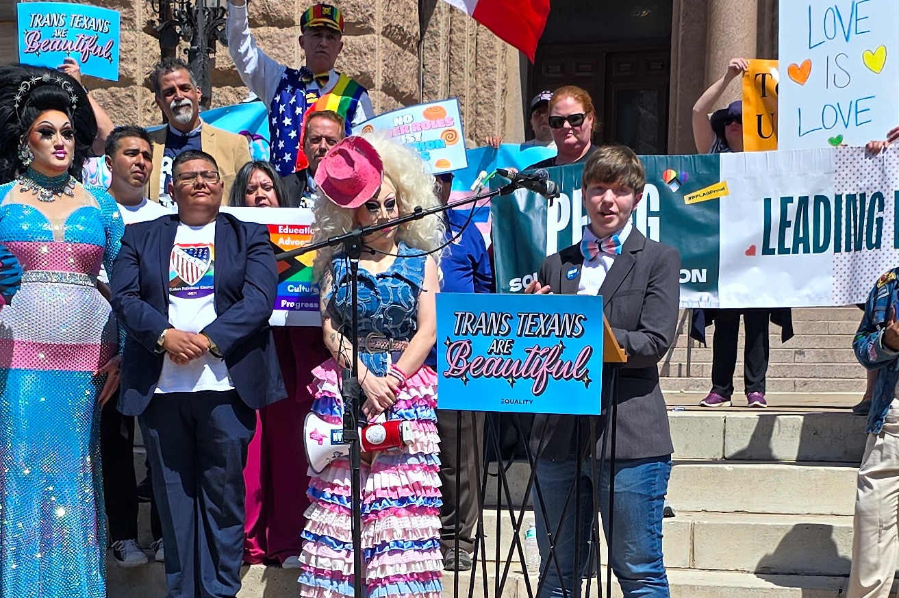 Ash Hall is a nonbinary white person with short brown hair in a pageboy cut, wearing a suit with a trans pride bowtie, speaks at a podium labeled Trans Texans are Beautiful. Other allies are arrayed behind them including colorful drag queens like Brigitte Bandit and Maxine LaQueen.