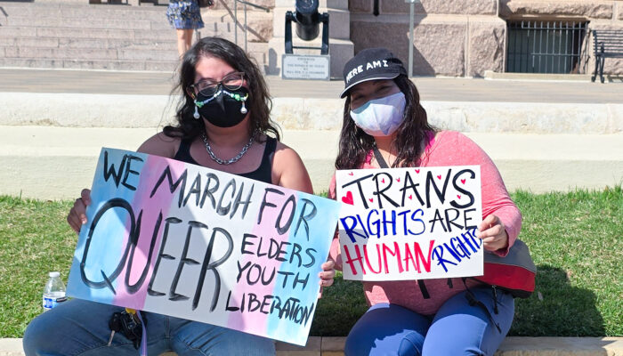 Two people with long dark hair pose with pro-trans signs: We March for Queer Elders/Youth/Liberation and Trans Rights Are Human Rights. Both people are masked, with the person on the left wearing a mask that is decorated with an ornate beaded chain.