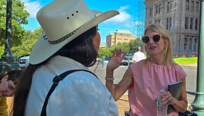 Former State Senator Wendy Davis, wearing sunglasses and a pink sleeveless top, holding a notebook and bottle water, waves a hand as she speaks with Sadie Hernandez of the Transgender Education Network of Texas, at the Texas State Capitol. Sadie is wearing a white cowboy hat and jacket.