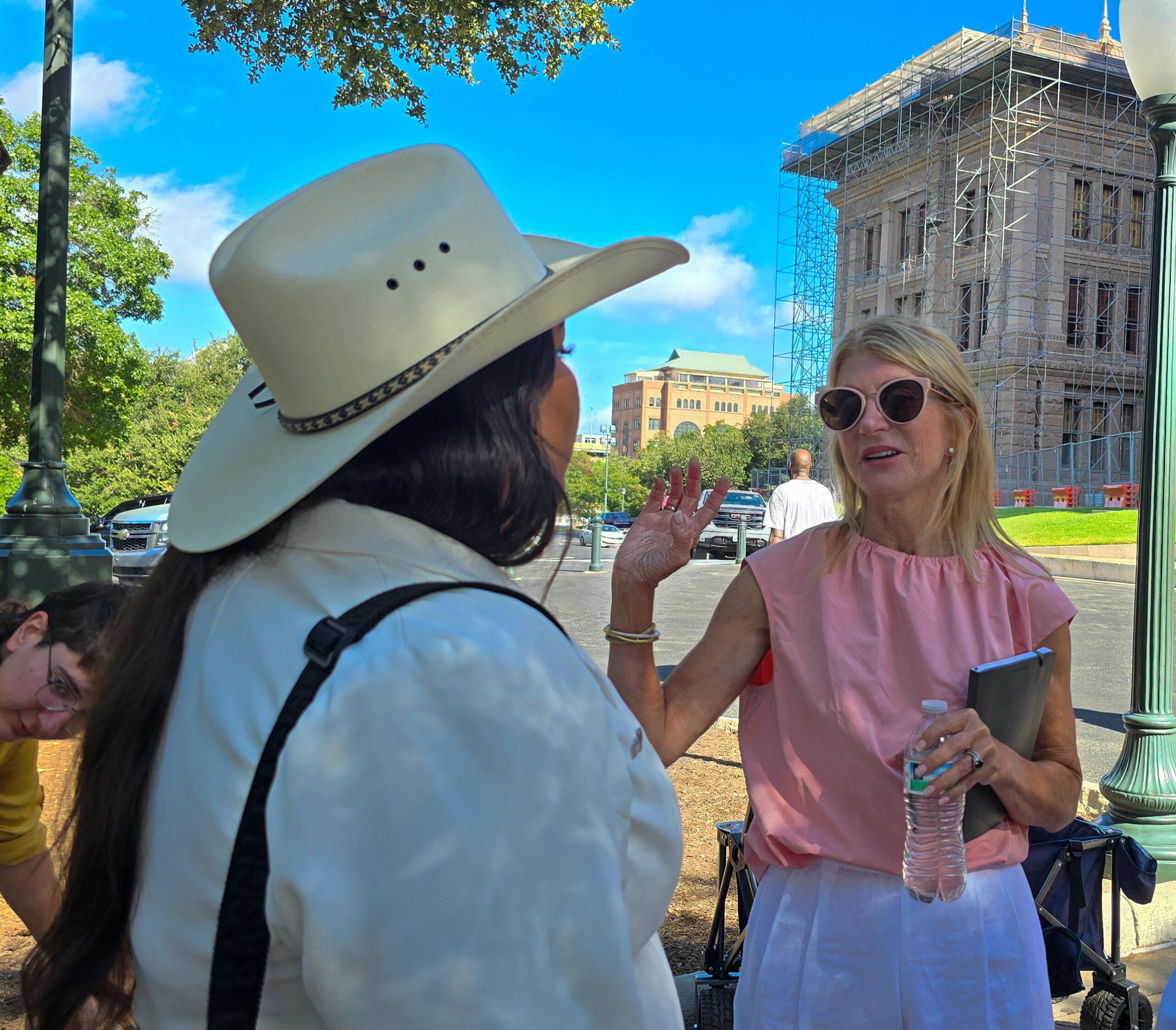 Former State Senator Wendy Davis, wearing sunglasses and a pink sleeveless top, holding a notebook and bottle water, waves a hand as she speaks with Sadie Hernandez of the Transgender Education Network of Texas, at the Texas State Capitol. Sadie is wearing a white cowboy hat and jacket.