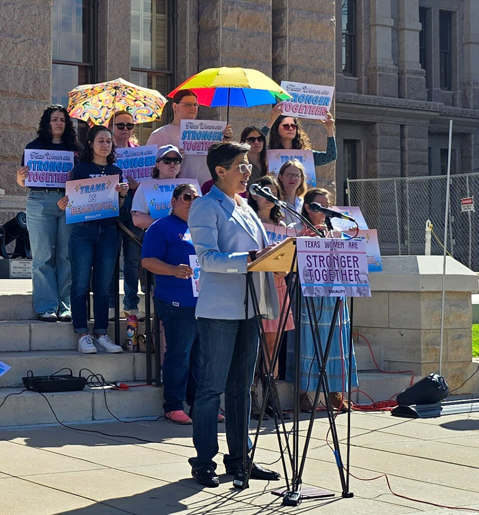 State Representative Jessica Gonzalez speaks at a podium labeled Texas Women Are Stronger Together, at the Texas Capitol south steps. She's wearing a light blue suit jacket and dark blue pants. Behind her, a group of mostly cisgender women show their support for trans people.
