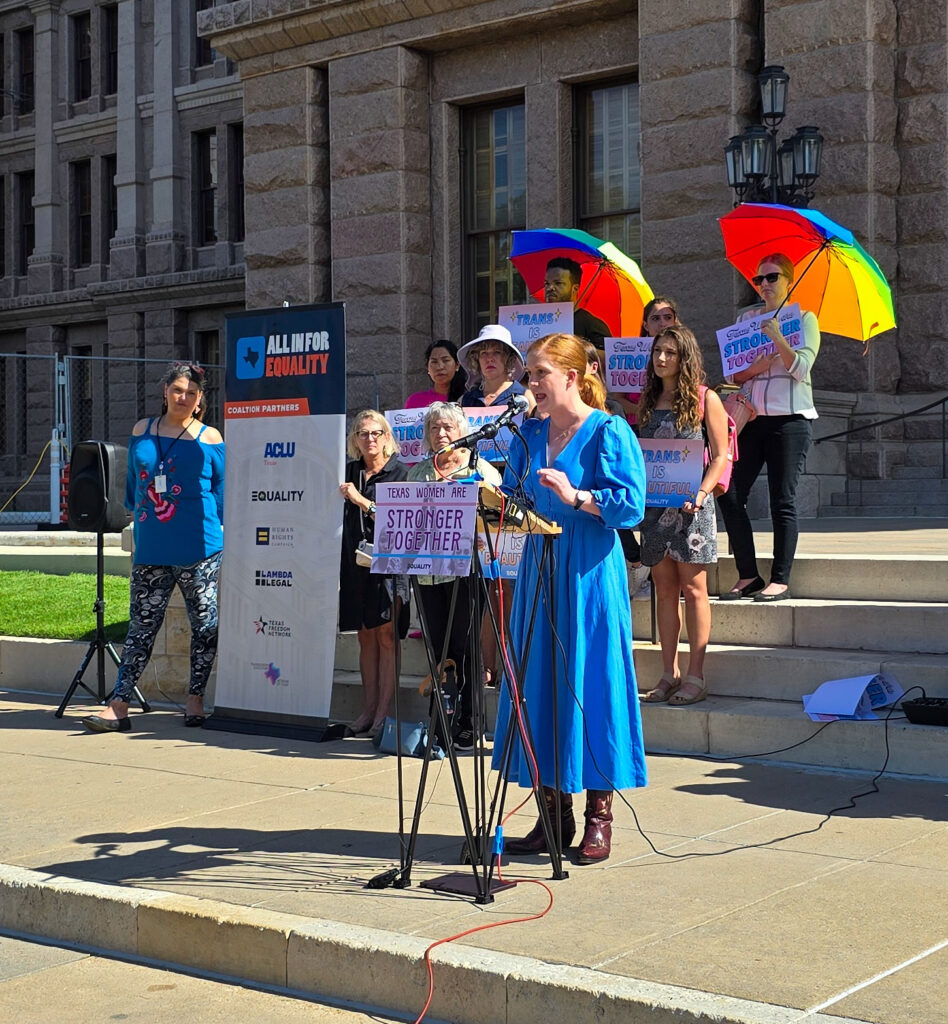 State Senator Molly Cook gestures with her hands as she speaks at the Texas Women Are Stronger Together podium at the Texas Capitol, wearing a blue dress and brown cowboy boots.