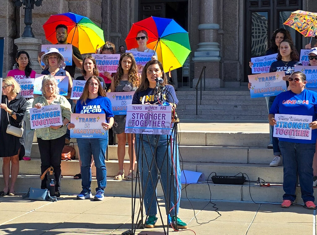 Ana Gonzalez is wearing blue jeans and "Union" sweatshirt as she speaks at a podium, surrounded by a group of mostly cisgender women showing their support for transgender rights.