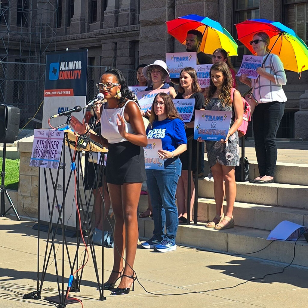 Kimiya Factory gestures with her hands as she speaks at a podium at the Texas Capitol, during a rally for transgender rights. She's dressed in patterned glasses, high heels, a black skirt, and white tank top.